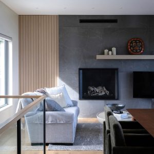 A modern living room with a grey sofa, cushions, and a blanket next to a window. The wall features wood and dark stone, a built-in fireplace, a floating shelf with candles and decor, and a glass railing.