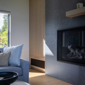A modern living room corner with a gray sofa, light wood flooring, a black-framed fireplace, and a large window letting in natural light. A round coffee table and textured rug are also visible.