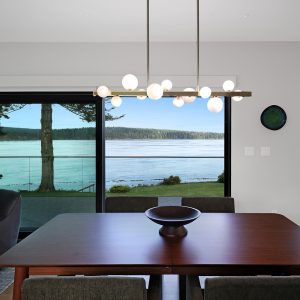 A modern dining room with a wooden table and chandelier overlooks a scenic lake view through large glass sliding doors. Trees and water are visible outside, and part of a kitchen is seen on the right.
