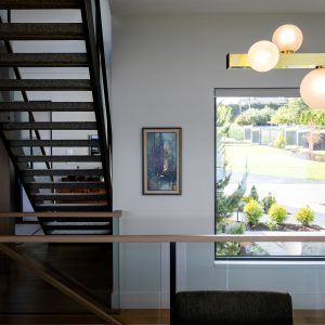 A modern interior with a wooden table, black chair, staircase with open risers, a framed painting on a white wall, a large window showing greenery outside, and a gold light fixture with round bulbs.