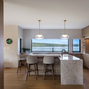 Modern kitchen with wooden cabinets, marble island, three bar stools, and pendant lights. Large windows offer a scenic view of a lake and forested hills. Dining area and living space are partially visible.