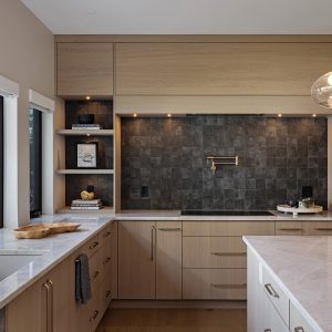 Modern kitchen with light wood cabinets, marble countertops, gold fixtures, a large window with an outdoor view, black tile backsplash, built-in shelves, and stainless steel oven appliances.