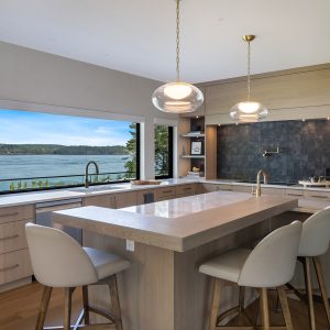 Modern kitchen with light wood cabinets, an island with seating, pendant lights, built-in appliances, and a large window offering a scenic view of a lake and trees outside.