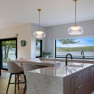 Modern kitchen with marble island, two pendant lights, and barstools. Large windows showcase a scenic view of water and trees, bringing in natural light. Light wood floors and minimal decor create an airy, open feel.