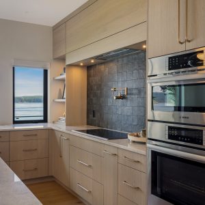 Modern kitchen with light wood cabinets, a marble countertop, built-in stainless steel ovens, a black tile backsplash, open shelving, and a window with a view of water and trees.