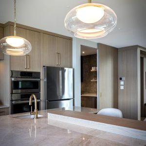 Modern kitchen with light wood cabinets, stainless steel appliances, marble countertop, and gold fixtures. Two round pendant lights hang above the island, creating a clean and contemporary atmosphere.