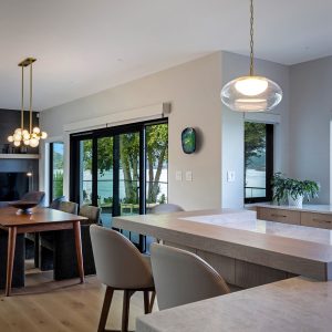 Modern kitchen and dining area with light wood floors, a marble island with beige barstools, pendant lights, a wooden dining table, dark chairs, and large windows overlooking trees and a green backyard.