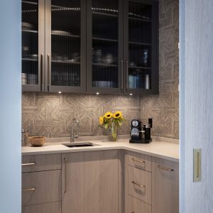 Modern kitchen with light wood cabinets, dark glass-front upper cabinets, geometric patterned backsplash, and built-in lighting. A vase of sunflowers and a coffee maker are on the countertop near a small sink.