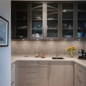 A modern kitchen with light wood cabinets, dark glass upper cabinets, geometric tile backsplash, a sink, sunflowers in a vase, a coffee machine, and wooden flooring under bright ceiling lights.