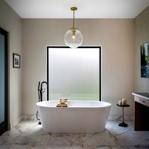A modern bathroom with a freestanding white bathtub, a gold light fixture above, marble floor tiles, a large frosted window, a small side table with candles, and minimalist decor.