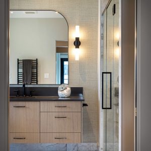 Modern bathroom with light wood vanity, black fixtures, large mirror, wall sconce, marble floor, glass shower door, towel rack with folded towel, and decorative sculpture on the counter. Neutral and sleek design.