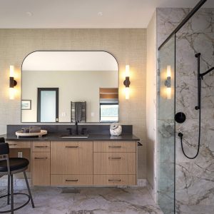 Modern bathroom with a floating wood vanity, black fixtures, decorative lighting, a large mirror, a glass-enclosed marble shower, a black chair, and a landscape painting on the wall. The room has a neutral color palette.