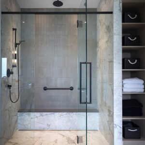 Modern bathroom with a glass-enclosed shower, marble walls and floor, a black rainfall showerhead, a built-in bench, grab bar, and open shelving with black baskets and neatly folded white towels.