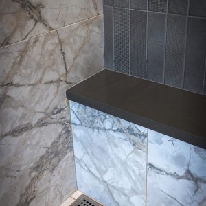 A modern bathroom corner with marble-patterned tiles on the walls and floor, a dark countertop, and a linear floor drain. The design features both large marble tiles and small rectangular textured tiles.