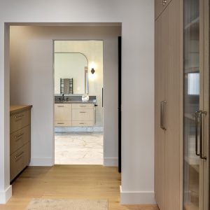 A modern walk-in closet with wooden cabinets and drawers on both sides, leading to a bathroom with light wood cabinetry, a large mirror, wall sconces, and a stone floor.