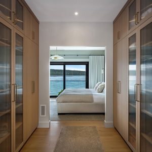 A modern walk-in closet with wooden cabinets and glass doors leads to a bedroom with a large bed and a window offering a scenic view of water and trees outside.