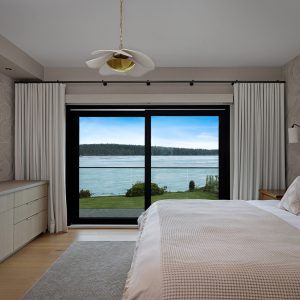 Modern bedroom with a large bed, light bedding, and decorative pillows, facing floor-to-ceiling windows with a scenic view of a river or lake, green landscape, and distant forested hills. Light décor and natural lighting fill the room.