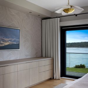 A modern bedroom with light wood furniture, a large window with white curtains overlooking a lake and forest, neutral walls with a leaf pattern, and a ceiling light shaped like a flower.
