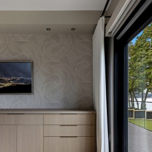 Modern bedroom corner with light wood cabinets, abstract patterned wallpaper, a wall-mounted TV, and large glass doors opening to a view of trees and a lake outside. White curtains are partially drawn to one side.