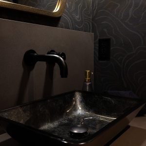 A modern bathroom features a rectangular black sink, matte black wall-mounted faucet, soap dispenser, and a decorative mirror. The wall has a dark, wavy, gold-accented pattern.