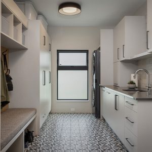 Modern laundry room with patterned tile floor, white cabinets, gray countertop, stacked washer and dryer, built-in bench with hooks and storage, and a frosted window letting in natural light.