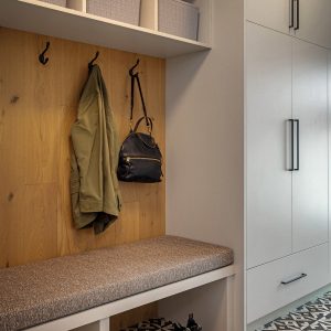 A mudroom with a patterned tile floor, built-in bench with cushion, cubby baskets above, two hooks holding a green jacket and black bag, and shoes and slippers placed neatly underneath.
