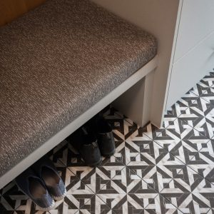 A patterned tile floor with black and white geometric designs, a cushioned bench, and two pairs of shoes—one blue and one black—placed neatly underneath the bench.