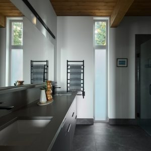 A modern bathroom with a dark countertop, black faucet, rectangular sink, large mirror, towel warmer, tall narrow windows, and a glass shower enclosure. The space has minimal decor and exposed wooden ceiling beams.