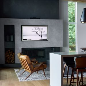 A modern living room with wooden floors, two brown chairs facing a fireplace, a monochrome tree photo above the mantel, a stack of firewood, and large windows letting in natural light.