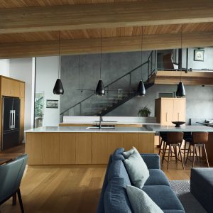Modern open-concept kitchen and living area with wood cabinets, a large island, blue sectional sofa, black pendant lights, dining table, and a staircase leading to a loft above. High ceiling with exposed wooden beams.