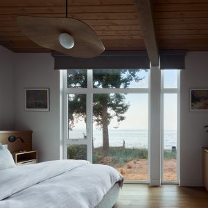 A modern bedroom with a large window overlooking trees and a view of the ocean. The room has a wooden ceiling, white walls, a neatly made bed, a dresser, and wall art. Natural light fills the space.