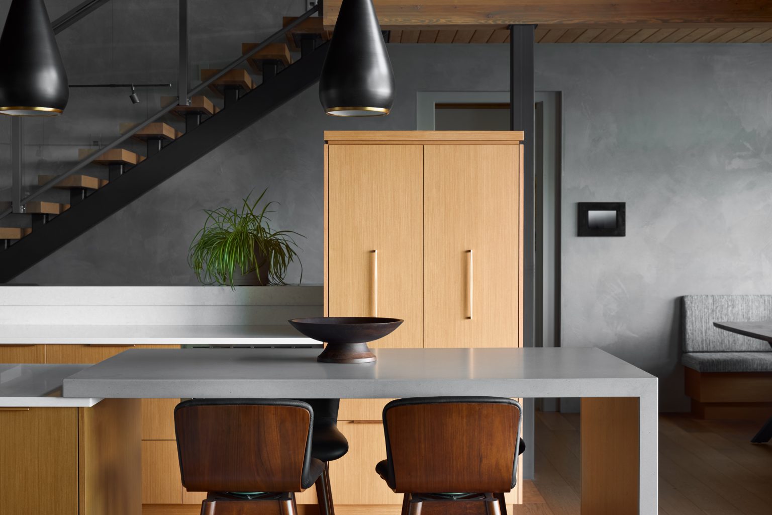 A modern kitchen with light wood cabinets, a gray countertop island, two dark wood chairs, a decorative bowl, two black pendant lights, and a green plant. A staircase and a gray wall are in the background.