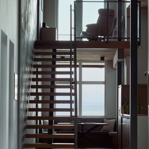 Modern interior with a wooden floating staircase, glass railing, and large windows letting in natural light. Upstairs, a lounge chair faces the windows, with a view of the ocean in the background.