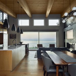 Modern open-concept kitchen and dining area with wood ceilings, large windows overlooking a body of water, a kitchen island with pendant lights, and a dining table with gray chairs. Contemporary decor and abundant natural light.