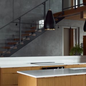 Modern kitchen with light wood cabinets, white countertops, black pendant lights, a built-in stove, and a black faucet. In the background, there is a staircase with glass railing and an upper mezzanine.