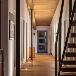 A modern hallway with wooden floors and ceiling, white walls, recessed lighting, framed art, and a staircase with wooden steps and metal railings leading up on the right. A glass-paneled door is at the end of the hallway.