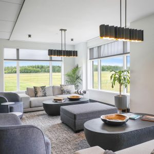 A modern living room with large windows showing a grassy outdoor view, grey sofas and chairs, black coffee tables with wooden bowls, two rectangular pendant lights, a plant, and books on the table.