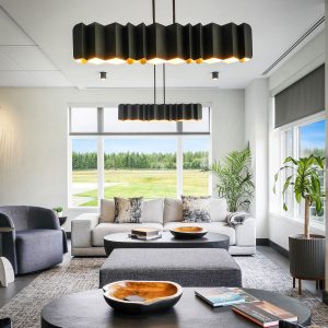 Modern living room with large windows, gray sofas, black coffee tables, wooden bowls, and indoor plants. Contemporary pendant lights hang from the ceiling, and a scenic green landscape is visible outside.