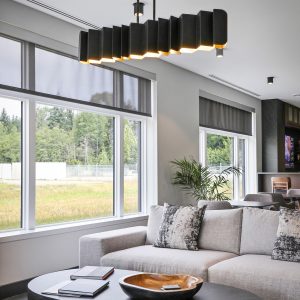 Modern living room with large windows, a light gray sectional sofa, patterned pillows, a black oval coffee table with a decorative bowl, books, and a unique geometric black chandelier overhead. Lush greenery is visible outside.