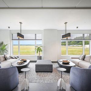 Modern living room with large windows overlooking a grassy field, featuring two light sofas, two dark armchairs, a central gray ottoman, a patterned rug, indoor plants, and contemporary pendant lights.