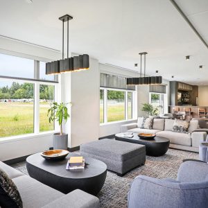 Modern lounge with large windows overlooking a grassy field, featuring gray sofas, black coffee tables, pendant lights, a dining area, and a bar in the background. Natural light fills the spacious, comfortable room.