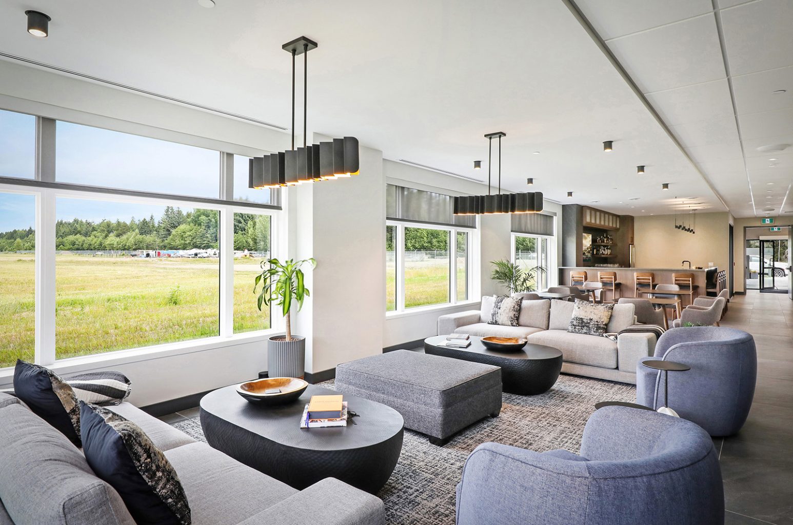 A modern lounge with large windows, gray sofas, black coffee tables, and pendant lights. The space has a dining area and bar in the background, with a view of a grassy field and trees outside.