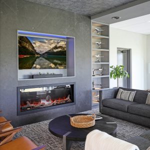 A modern living room with a gray sofa, round black coffee table, and tan chairs. A built-in electric fireplace sits below a TV displaying a lake scene. Shelves with model airplanes are on the right wall.