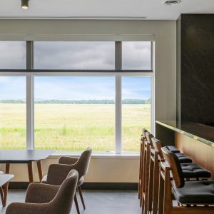 A modern lounge area with upholstered armchairs and tall wooden barstools sits by large windows overlooking a green field under a cloudy sky.