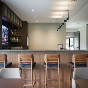 A modern bar area with wooden stools and chairs, a TV showing a news channel on the left, a small bar counter with shelves of bottles, and minimalist lighting hanging from the ceiling.