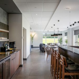 Modern lounge with a sleek kitchen area, wooden bar stools, coffee station, and seating area with armchairs. Large windows let in natural light and offer views of greenery outside.