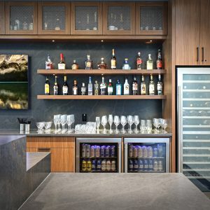 A modern home bar with liquor bottles on wooden shelves, a wine fridge, two mini fridges with drinks, glassware, and a wall-mounted TV showing a scenic lake and mountain landscape.