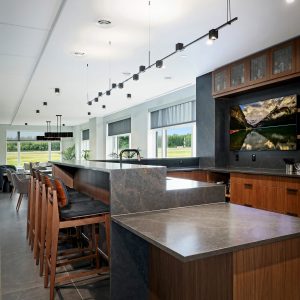 Modern kitchen with sleek dark countertops, wood cabinetry, bar stools, pendant lighting, and large windows showing a green outdoor view. A digital screen above the counter displays a scenic mountain landscape.