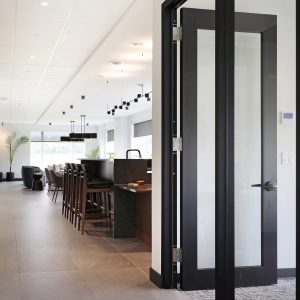 A modern open-plan kitchen and dining area with sleek black-framed glass doors, high bar stools, pendant lights, and large windows letting in natural light. The space features minimalist decor and neutral colors.