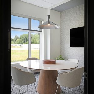 A modern small meeting room with a round table, four white chairs, a large window showing greenery outside, a pendant light, a wall-mounted TV, and a plant in the corner.